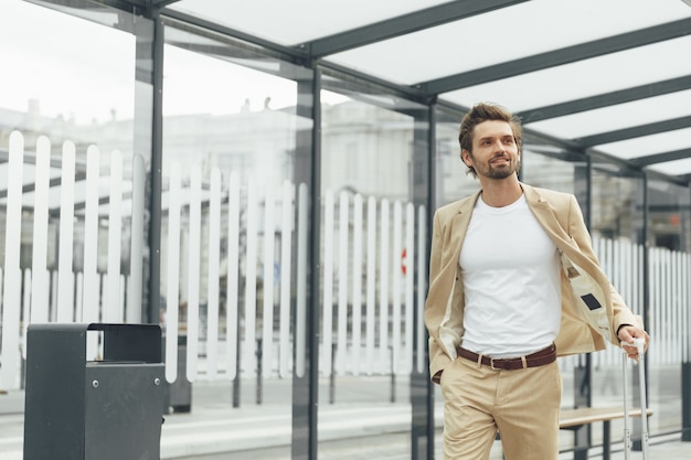 Hombre guapo con barba en ropa formal caminando en la estación de autobuses con maleta. Joven empresario con viaje de trabajo en otra ciudad