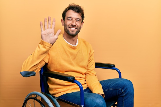 Hombre guapo con barba sentado en silla de ruedas mostrando y señalando con los dedos número cinco mientras sonríe confiado y feliz