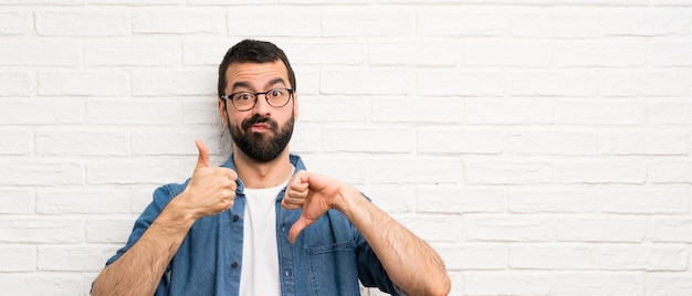 Hombre guapo con barba sobre pared de ladrillo blanco haciendo señal de bueno a malo. Indecisa entre sí o no