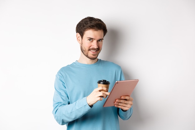 Hombre guapo con barba sonriendo a la cámara, tomando café y leyendo en tableta digital, de pie sobre fondo blanco. Copia espacio