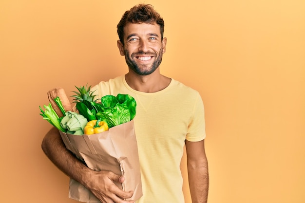 Hombre guapo con barba sosteniendo una bolsa de papel con pan y comestibles con aspecto positivo y feliz de pie y sonriendo con una sonrisa segura mostrando los dientes