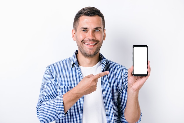 Hombre guapo con barba sosteniendo smartphone y apuntando con el dedo a la pantalla mientras mira a la cámara. Foto de estudio con fondo blanco. Concepto de personas y tecnologías