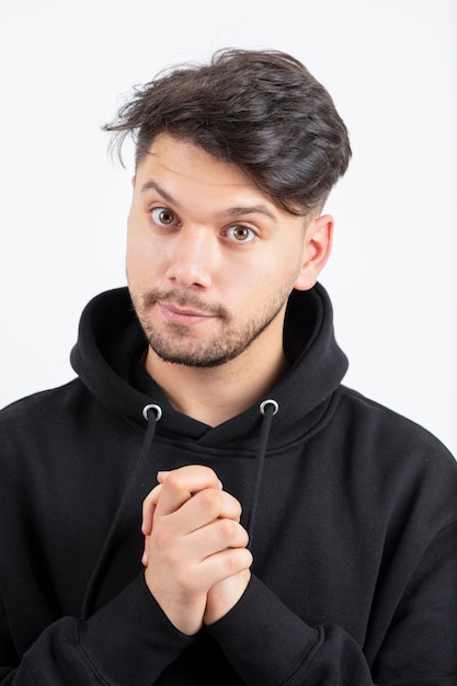 Hombre guapo con barba en sudadera con capucha negra de pie sobre la pared blanca