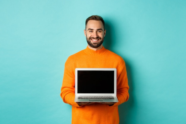 Hombre guapo con barba en suéter naranja que muestra la pantalla del portátil, demostrando tienda en línea, de pie sobre fondo azul claro.