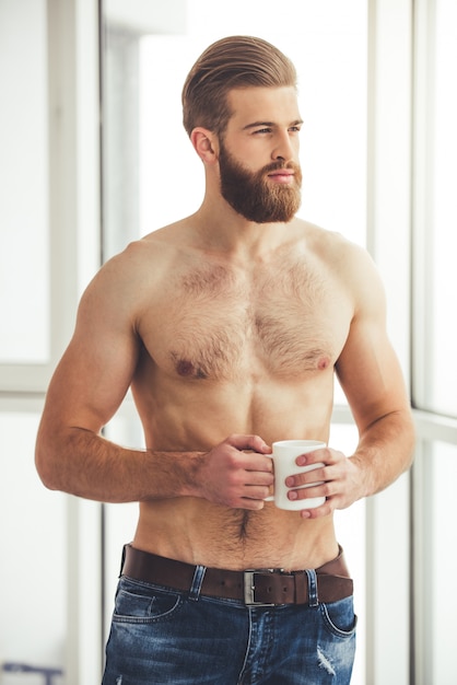 Hombre guapo con barba y el torso desnudo está sosteniendo una taza.