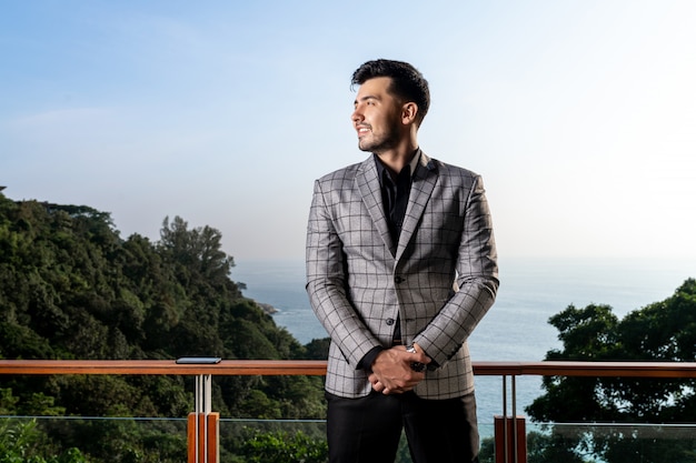 Hombre guapo con barba en un traje formal se encuentra en un balcón con una hermosa vista de la naturaleza