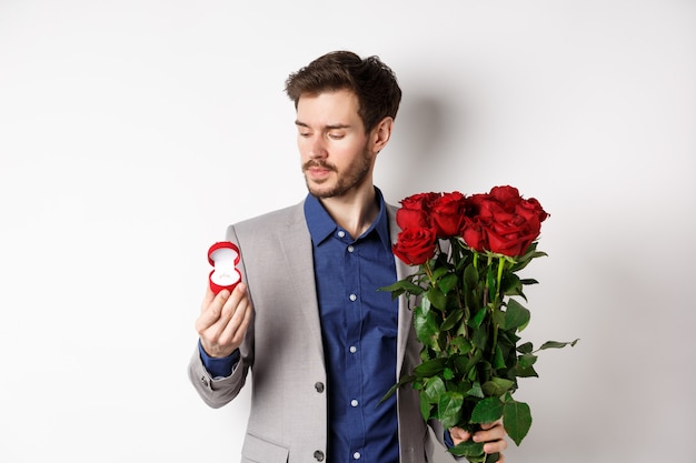 Hombre guapo con barba en traje mirando el anillo de compromiso, haciendo sorpresa en el día de los enamorados, de pie con rosas rojas sobre fondo blanco.