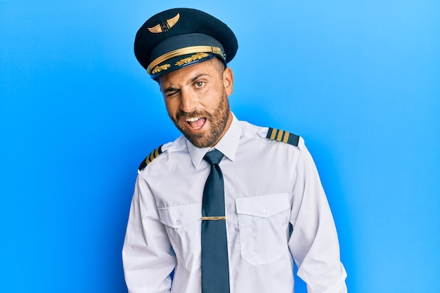 Hombre guapo con barba con uniforme de piloto de avión guiñando un ojo mirando a la cámara con expresión sexy, cara alegre y feliz.