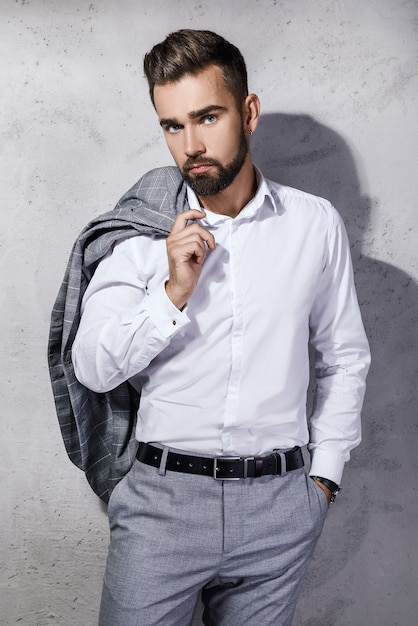 Hombre guapo con barba vestido con traje gris está posando contra la pared de hormigón