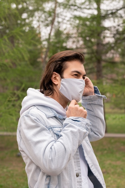 Hombre guapo con cabello castaño caminando en el parque y ajustando la máscara de tela en la cara durante la epidemia de coronavirus