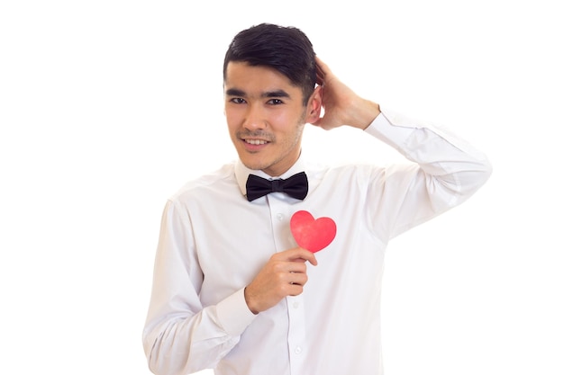 Hombre guapo con cabello negro en camiseta blanca con pajarita negra sosteniendo un corazón de papel rojo sobre fondo blanco en el estudio