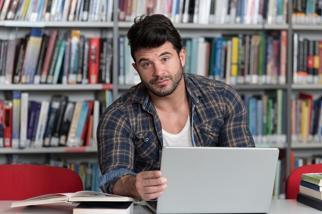 Hombre guapo con cabello oscuro sentado en un escritorio en la computadora portátil de la biblioteca y organizador en la mesa mirando la pantalla un concepto de estudiar libros borrosos en la parte posterior