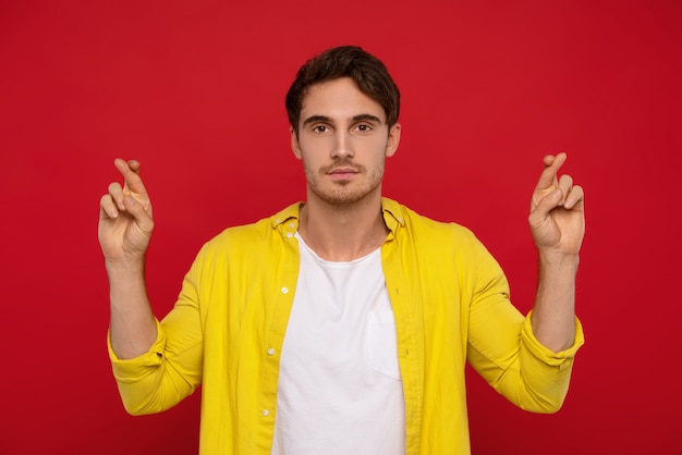 Hombre guapo en camisa amarilla con los dedos cruzados aislado sobre fondo rojo.