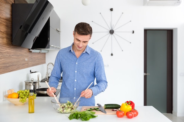 Hombre guapo en camisa azul de pie y mezcla de verduras para ensalada en la cocina