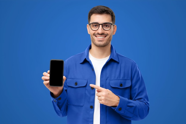 Foto hombre guapo con camisa azul presentando teléfono inteligente y señalando con el dedo la pantalla negra en blanco