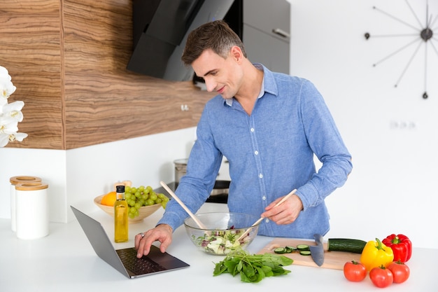 Hombre guapo con camisa azul usando laptop y cocinando en la cocina de casa