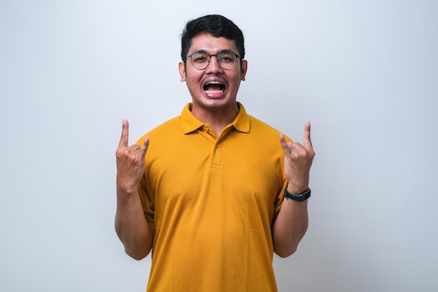 Hombre guapo con camisa casual sobre fondo blanco gritando con expresión loca haciendo símbolo de rock con las manos arriba Estrella de la música Concepto pesado