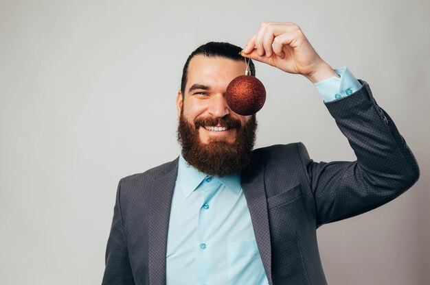 Foto un hombre guapo con camisa y chaqueta cubre un ojo con un globo navideño rojo. estudio grabado sobre fondo gris. anuncio de fiesta de navidad en la oficina.