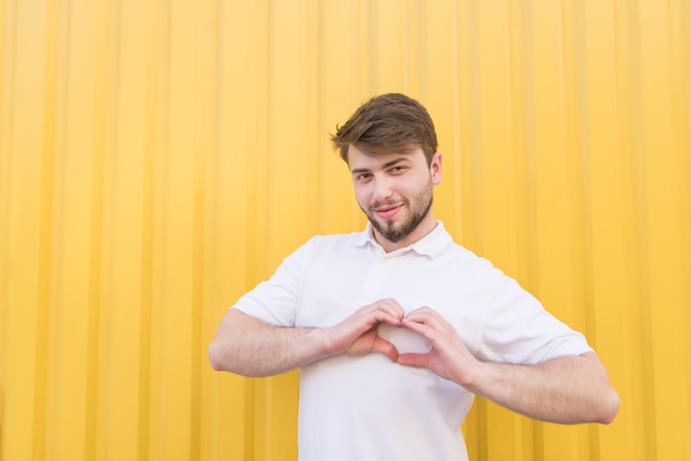 Un hombre guapo con una camiseta blanca y una barba muestra una mano con un signo de corazón en una pared amarilla.