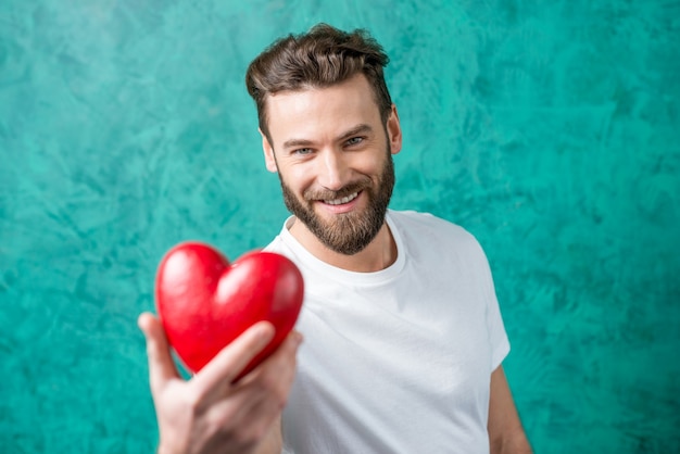 Foto hombre guapo en la camiseta blanca dando pie de corazón rojo sobre el fondo de la pared pintada de verde. concepto de san valentín