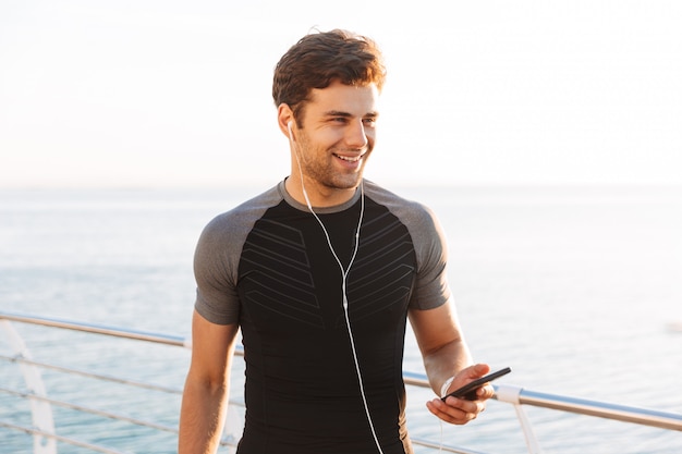 Hombre guapo en camiseta escuchando música a través de auriculares inalámbricos, durante la caminata al aire libre en la playa