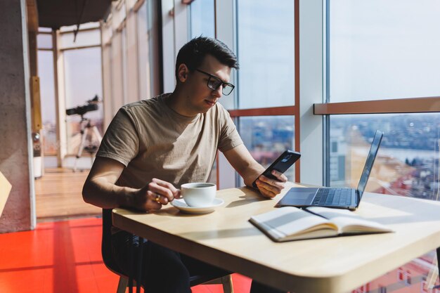 Un hombre guapo con una camiseta informal roja escribiendo un mensaje en el teléfono móvil mientras se sienta cerca de una ventana de vidrio en la mesa con una computadora portátil que trabaja de forma remota en un café