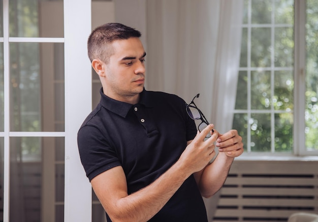 un hombre guapo con una camiseta negra considera anteojos elegantes con un borde negro en las manos en la habitación al fondo de la ventana. De cerca