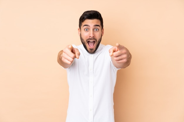 Hombre guapo caucásico aislado en pared beige sorprendido y apuntando al frente