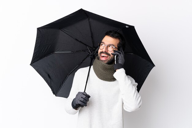 Hombre guapo caucásico con barba sosteniendo un paraguas sobre una pared blanca aislada manteniendo una conversación con el teléfono móvil