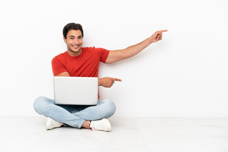 Foto hombre guapo caucásico con una computadora portátil sentada en el suelo apuntando con el dedo hacia un lado y presentando un producto