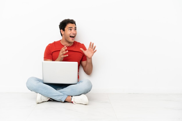 Hombre guapo caucásico con una computadora portátil sentada en el suelo con expresión facial sorpresa