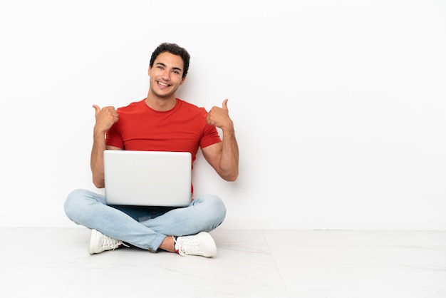 Hombre guapo caucásico con una computadora portátil sentada en el suelo con los pulgares hacia arriba gesto y sonriendo