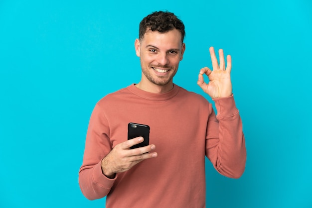 Hombre guapo caucásico joven aislado en azul con teléfono móvil y haciendo el signo de OK