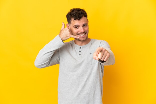 Hombre guapo caucásico joven aislado en la pared amarilla haciendo gesto de teléfono y apuntando hacia el frente