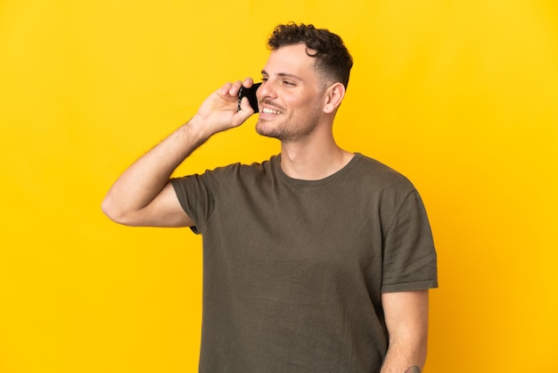 Hombre guapo caucásico joven aislado en la pared amarilla manteniendo una conversación con el teléfono móvil