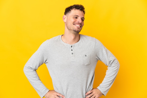 Hombre guapo caucásico joven aislado en la pared amarilla posando con los brazos en la cadera y sonriendo