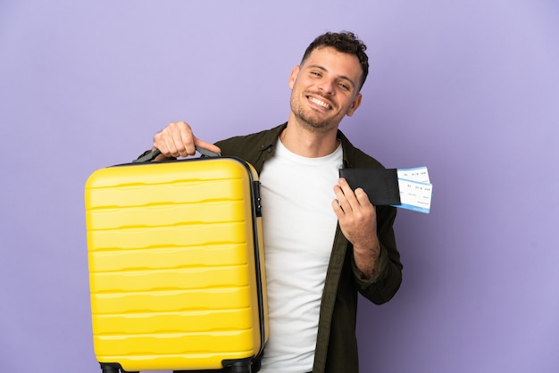 Hombre guapo caucásico joven aislado en la pared blanca en vacaciones con maleta y pasaporte