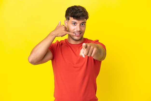 Hombre guapo caucásico joven aislado sobre fondo amarillo haciendo gesto de teléfono y apuntando hacia el frente