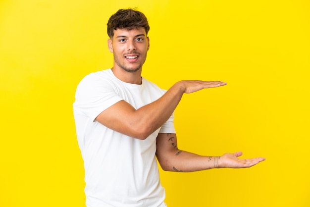 Hombre guapo caucásico joven aislado sobre fondo amarillo sosteniendo copyspace para insertar un anuncio