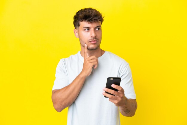 Hombre guapo caucásico joven aislado sobre fondo amarillo usando teléfono móvil y pensando