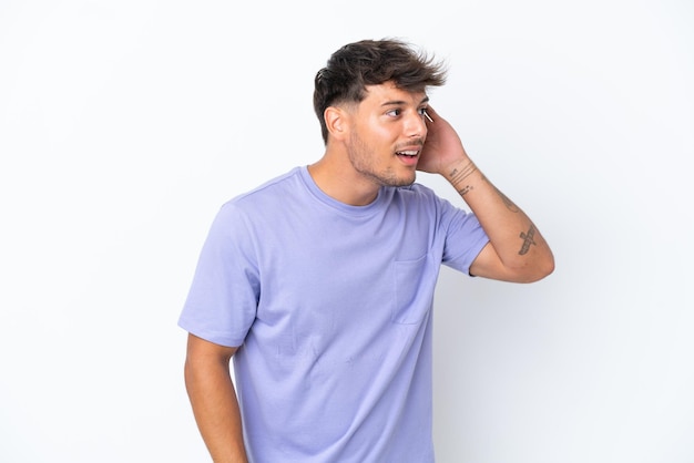 Hombre guapo caucásico joven aislado sobre fondo blanco escuchando algo poniendo la mano en la oreja