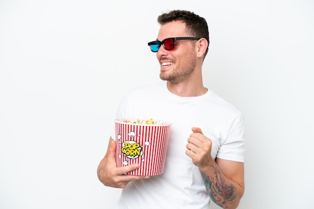 Hombre guapo caucásico joven aislado sobre fondo blanco con gafas 3d y sosteniendo un gran cubo de palomitas de maíz
