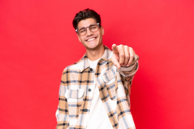 Hombre guapo caucásico joven aislado sobre fondo rojo apuntando al frente con expresión feliz