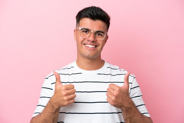 Hombre guapo caucásico joven aislado sobre fondo rosa con gafas y con el pulgar hacia arriba