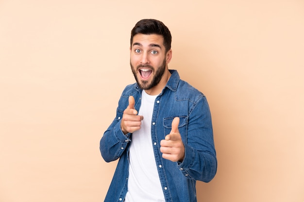 Hombre guapo caucásico en pared beige sorprendido y apuntando al frente