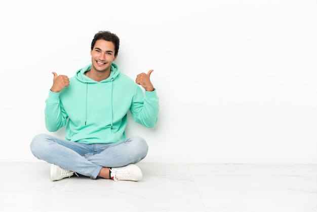 Hombre guapo caucásico sentado en el suelo con el pulgar hacia arriba gesto y sonriendo