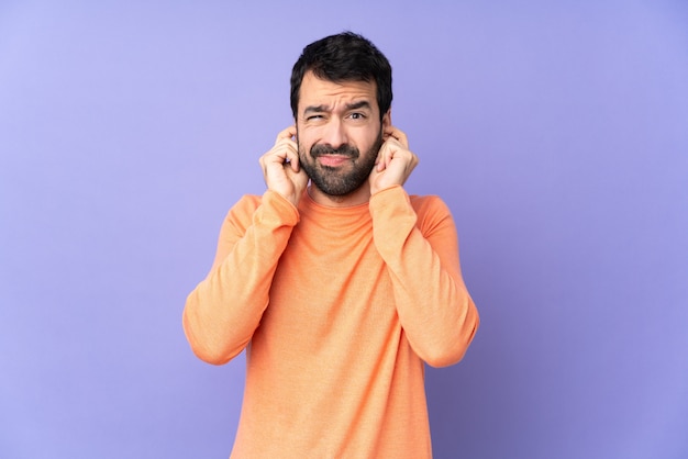 Hombre guapo caucásico sobre pared púrpura aislado frustrado y cubriendo las orejas