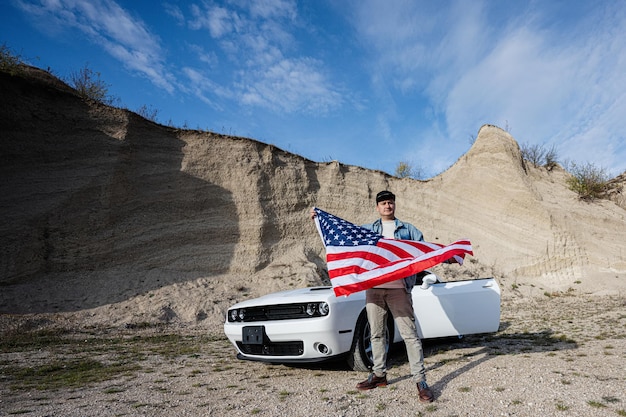 Hombre guapo con chaqueta de jeans y gorra con bandera de Estados Unidos cerca de su auto deportivo americano blanco en carrera