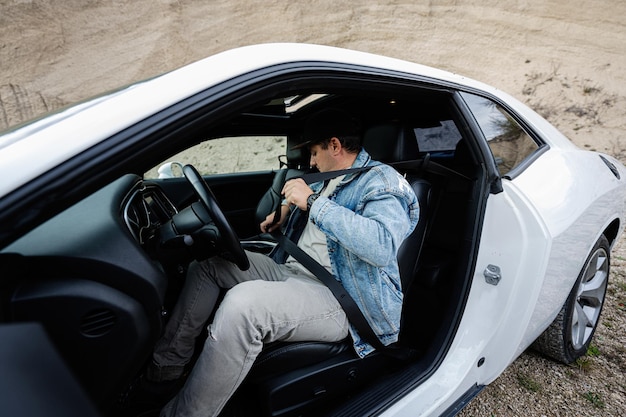 Un hombre guapo con chaqueta de jeans y gorra se sienta y se ata el cinturón en su auto deportivo blanco