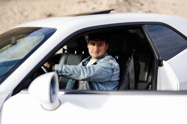 Un hombre guapo con chaqueta de jeans y gorra se sienta en su auto deportivo blanco